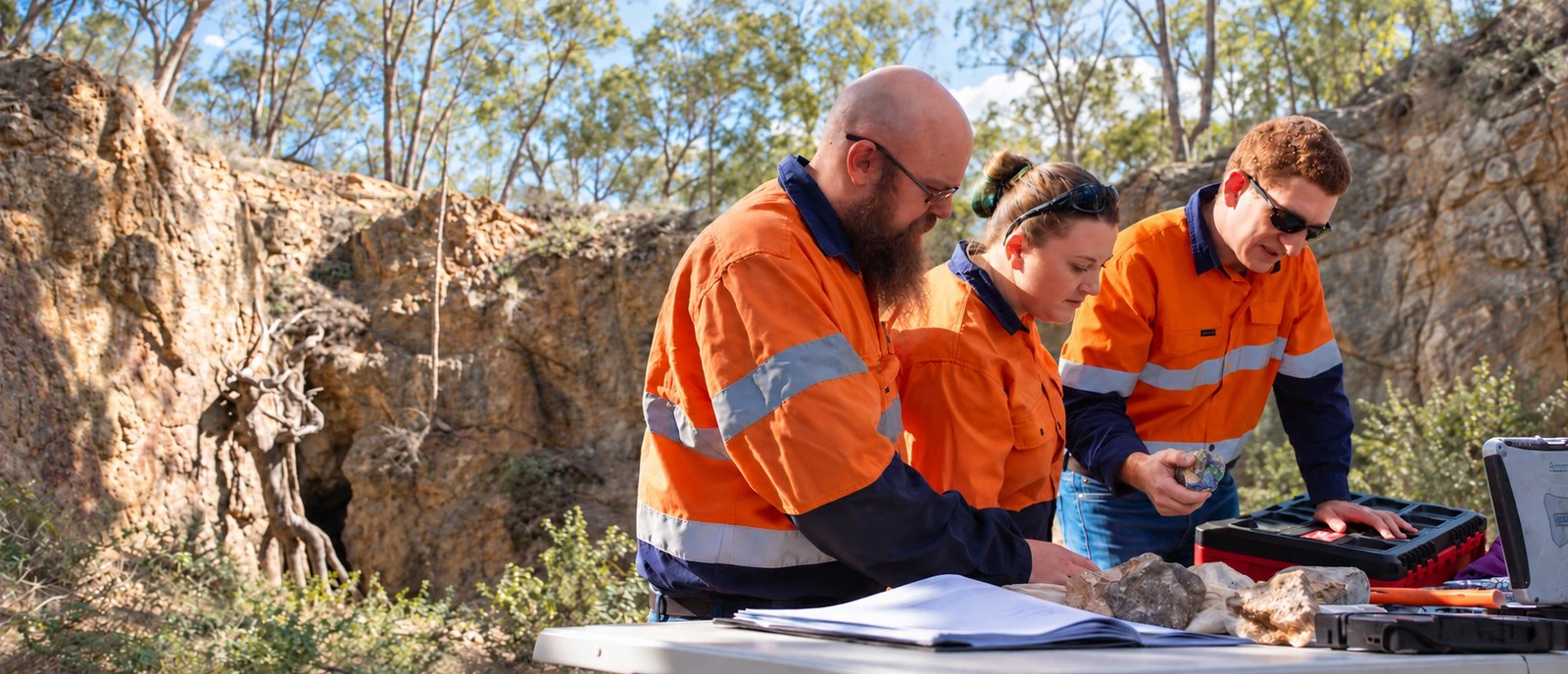 Exploration team examining a blue-and-green rock specimen and field records against a pit backdrop.